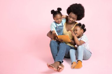 Aile okuma konsepti. İki sevimli Afrikalı kız renkli giysiler içinde güzel genç annelerinin onlara bir kitaptan hikayeler okumasını dinliyorlar. Pembe arkaplanda izole edilmiş, kopyalama alanı