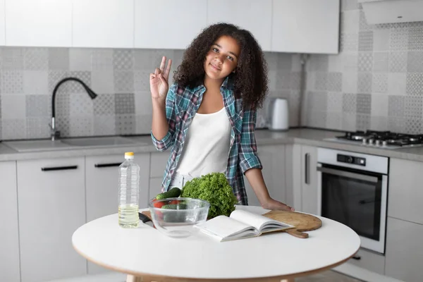 Portrait of mixed-race curly teen girl standing in the kitchen in front of a table with vegetables and a book for recipes, prepares a salad, looks at the camera and shows her hand a sign of peace. Stock Picture