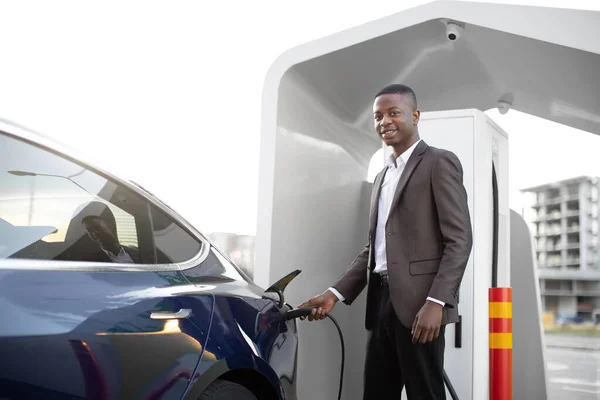 Voitures électriques, concept EV, carburant écologique. Portrait de jeune homme noir souriant en vêtements de travail, rechargeant sa voiture électrique de luxe moderne — Photo