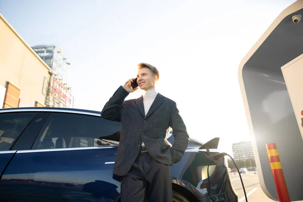 Véhicules et concept écologique. Occupé agréable confiant jeune homme européen charge sa voiture électrique le matin, avoir une conversation par téléphone — Photo