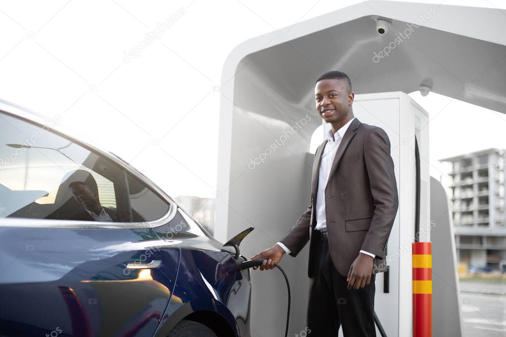 Electric cars, EV concept, eco friendly fuel. Portrait of young smiling black man in business clothes, recharging his modern luxury electric car