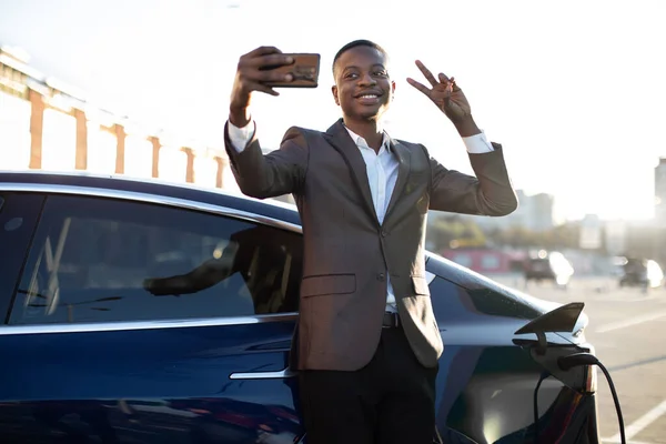 Concept de station-service électrique. Portrait de l'homme noir sourire confiant agréable en tenue formelle, montrant geste de paix à sa caméra smartphone, en attendant le ravitaillement en carburant de la voiture — Photo