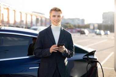 Yakışıklı, beyaz bir adam gülümsüyor, cep telefonuyla mesajlaşıyor ya da uygulamasını kullanıyor, açık hava benzin istasyonunda elektrikli araba şarj ederken.