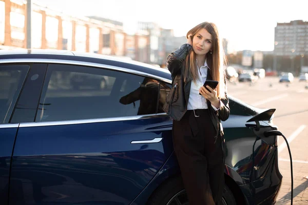 Jeune belle dame d'affaires réussie avec un long har, portant une tenue formelle noire et une chemise blanche, regarde la caméra en attendant son ravitaillement en carburant de voiture électrique — Photo