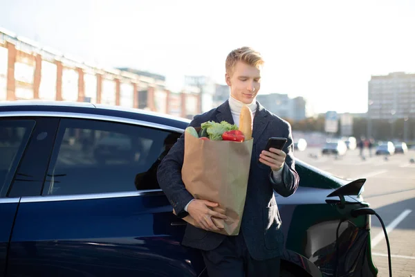 Beau jeune homme caucasien souriant, tenant sac à provisions avec des légumes, messagerie sur son téléphone portable ou en utilisant l'application pour payer, tout en chargeant la voiture électrique à l'extérieur à la station de ravitaillement de la ville — Photo