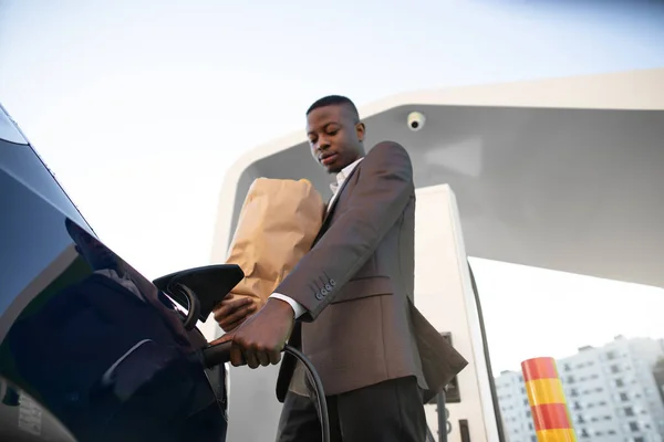 Vue du bas du jeune homme d'affaires africain en costume, rechargeant sa voiture électrique à la station de stationnement après avoir fait du shopping dans un centre commercial. Focus sur fiche de charge. — Photo