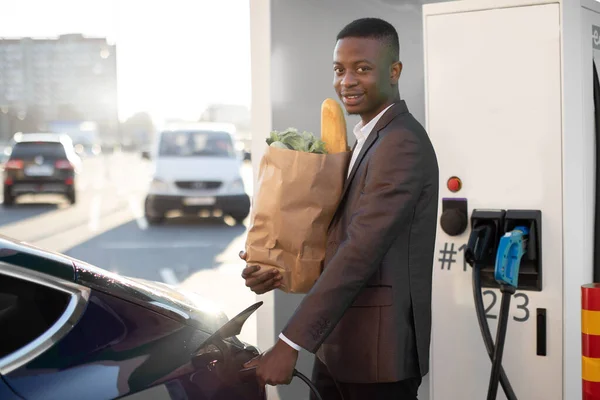 Portrait de jeune homme noir souriant en vêtements de travail, tenant un sac à provisions en papier avec de la nourriture, rechargeant sa voiture électrique de luxe moderne. Voitures électriques, concept EV, carburant écologique. — Photo