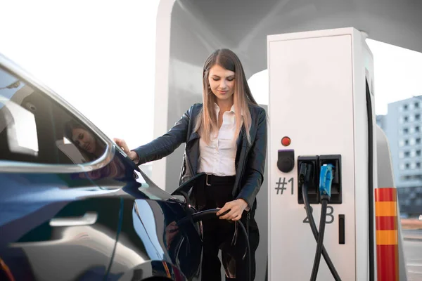 Borne de recharge EV, concept d'éco-carburant. Portrait de jolie jeune fille insérant fiche de charge dans sa voiture électrique moderne pour le ravitaillement à la gare de la ville — Photo