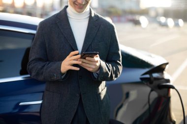 Şehrin şarj istasyonunda cep telefonuyla lüks elektrikli arabasının yanında duran yakışıklı delikanlının yakın plan çekimleri..