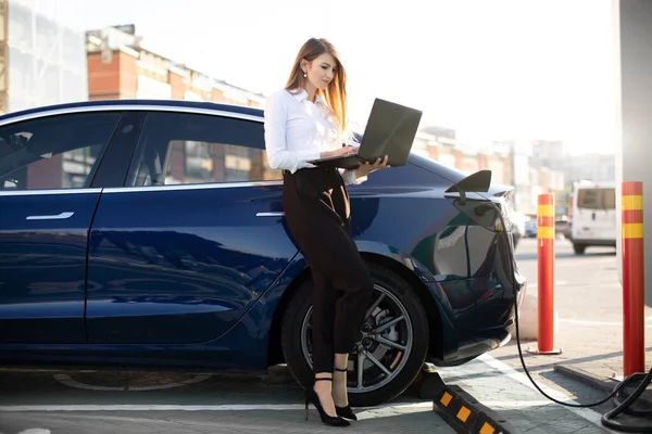 Jeune femme d'affaires en vêtements formels, debout avec un ordinateur portable près de sa voiture électrique éco, attendant la batterie de ravitaillement à la station de recharge extérieure de la ville — Photo