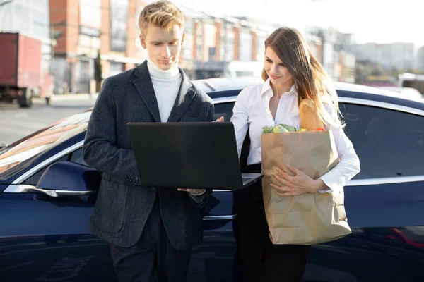 Lunch break, shopping of food, work everywhere. Handsome young man holds laptop pc and attractive woman with paper shopping bag with fresh food, standing near the modern car outdoors