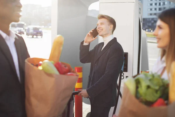 Blurred faces of happy young friends, black man and european woman, holding shopping bags with food and talking, while their busy handsome colleague charges electric car and talks phone