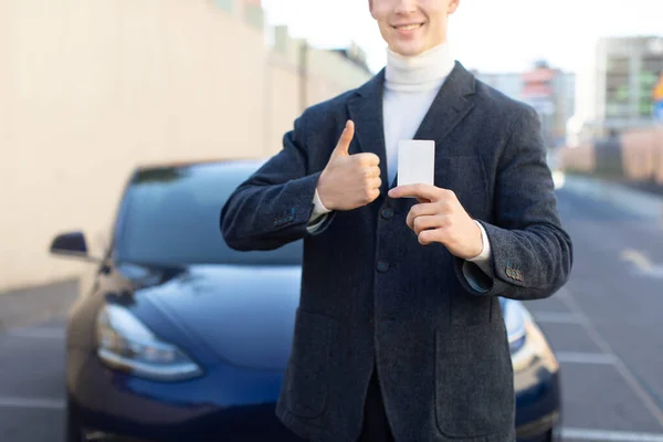 Concept de véhicule électrique futuriste. Coupé plan horizontal de sourire jeune homme caucasien concessionnaire automobile travailleur, détient carte clé de voiture électro moderne et montre pouce vers le haut — Photo