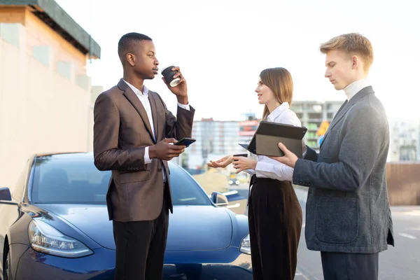 Groupe d'affaires multiethnique, deux hommes et une femme prenant une pause café, en utilisant une tablette et des smartphones et discuter du travail et du projet, debout près de la voiture électrique. Concept d'entreprise. — Photo