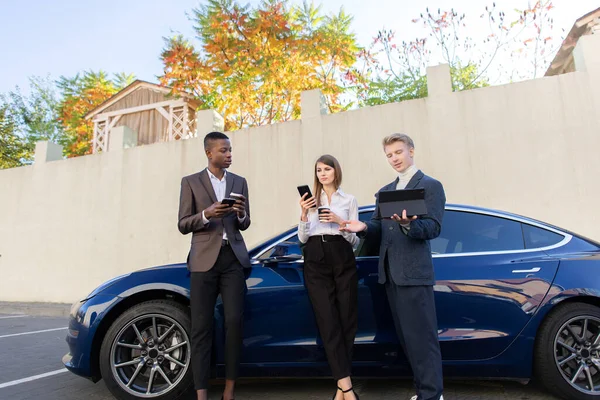 Les jeunes partenaires d'affaires de confiance, l'homme africain et l'homme et la femme caucasiens, travaillent en utilisant des tablettes et des téléphones ensemble avant le voyage d'affaires debout près de la voiture électro à l'extérieur. — Photo