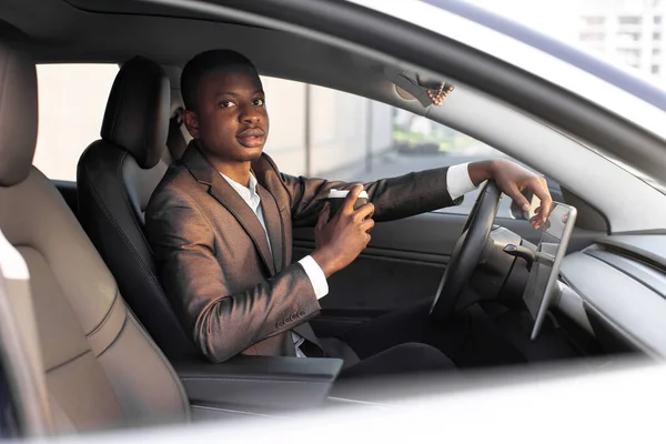 Handsome young dark skinned man in formal wear sitting in his modern electric car and is ready to start charging at EV charging station, while drinking coffee to go. Inside cabin view.