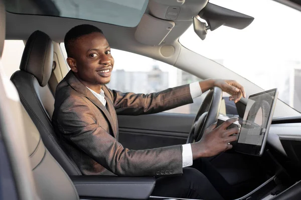Happy smiling African American male driver sitting behind the self-driving steering wheel of an autonomous electric modern car. Happy guy holds coffee cup and smiles to camera in modern electric car