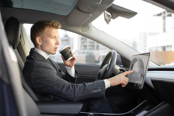Testing a new electric futuristic car with self driving system. Side view of handsome Caucasian businessman sitting in modern car, drinking coffee to go and touching navigation autopilot screen