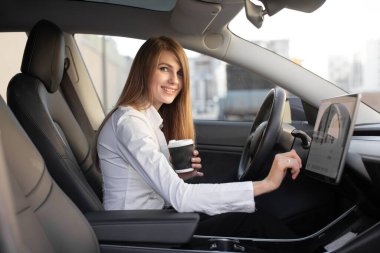 Beyaz tenli çekici iş kadını kahve içiyor modern lüks elektrikli arabalarda direksiyonda oturuyor ve navigasyon sisteminden geçmek için elektronik gösterge paneli kullanıyor.