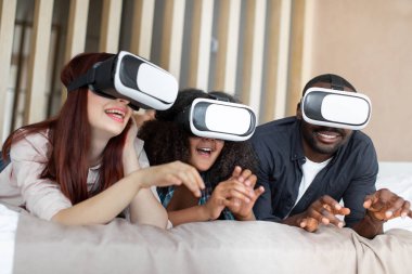 Gülümseyen çok ırklı bir aile, yatakta uzanıp film izlemek ya da sanal gerçeklik cihazında video oyunu oynamak.