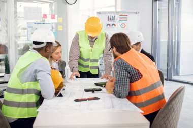 Çeşitli mimarlar ellerinde planlarla ofiste oturuyorlar.