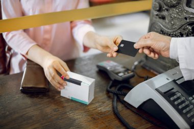 Erkek eczacının, tanınmayan bir kadın hastadan kredi kartı alırken, ilaç kutusunu tutarken, eczanedeki ilaçların parasını kredi kartıyla öderken çekilmiş görüntüsü..