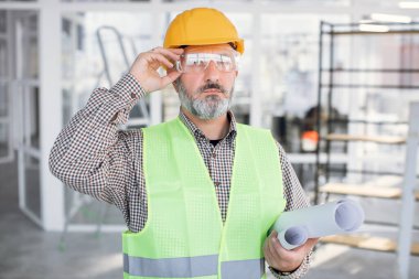 Gözlüklü ve miğferli yetenekli sakallı adam elinde planlarla poz veriyor.