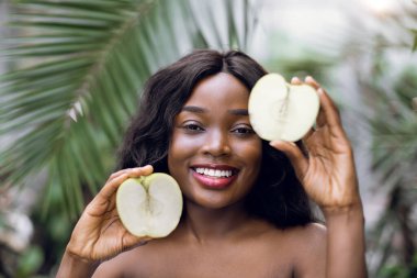 Mutlu, gülümseyen bir afro kadın seradaki palmiyelerin arasında elma yarımları arasında. Güzellik portresini kapat. Sağlıklı yaşam tarzı ve güzellik kavramı