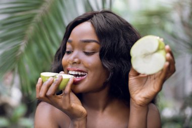 Tazelik, meyveler, insanlar ve yaz konsepti. Güzel Afro-Amerikan kadın modelinin yakın plan portresi, tropikal palmiye ve bitkilerin arka planında, elma parçalarıyla, elmanın yarısını ısıran