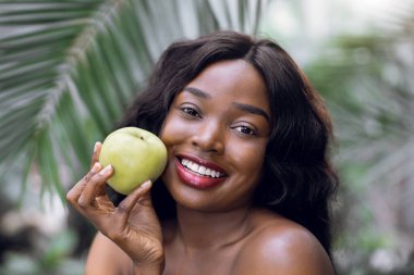 Esmer Afrikalı kadının çıplak omuzlu güzel portresi, yeşil elmalarla seranın arka planında poz veriyor. Sağlıklı meyve, yaz konsepti.