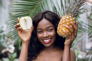 Sağlıklı gıda, vegan ve güzellik konsepti tanıtımı. Gülümseyen güzel Afrikalı kadın, mükemmel bir ten ve gülümsemeyle, palmiye yapraklarının arkasında poz veriyor ve elinde taze meyve, elma ve ananas tutuyor.