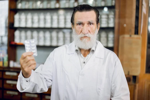 Take care of yourself, take medicine. Portrait of confident likable senior pharmacist in his ancient pharmacy, holding blister pack with white pills