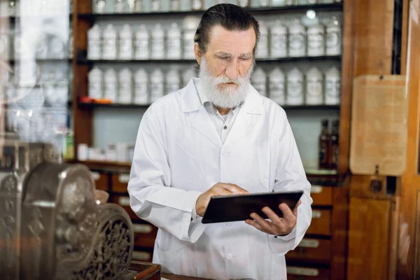 Portrait of concentrated high-skilled senior man pharmacist standing near vintage cash register at the counter in an old pharmacy and using tablet pc
