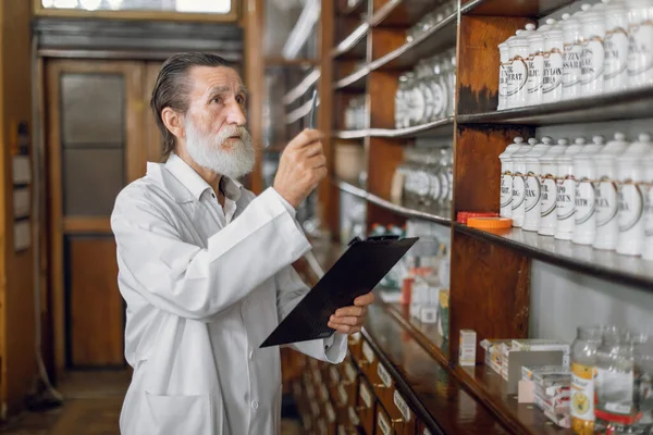 Drug control and inventory. Side view of pensive male pharmacist standing in vintage old drugstore, while using clipboard and pen for making medicines inspection