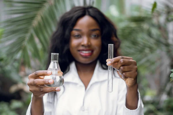 Yakın plan, serada deney tüpü ve yeşil bitkilerin önünde duran beyaz önlüklü, gülümseyen, tatlı Afrikalı kadın tarım uzmanının bulanık görüntüsü. — Stok fotoğraf