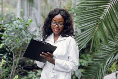 Bitki bakımı ve koruma konsepti. Serada çalışmak için. Şirin gülümseyen Afro-Amerikan kadın tarım mühendisi seradaki güzel palmiye ağaçlarının önünde duruyor ve notlar alıyor.