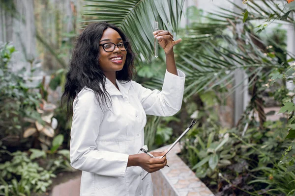 Biotechnology, agronomy, science and engineering concept. Young black female scientist holding test tube with sample and clipboard, smiling at camera, working in hothouse