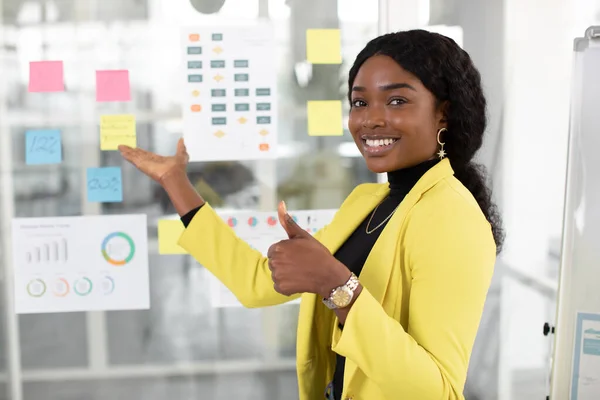 Ofiste poz veren parlak sarı ceketli, arka plandaki dosya ve çıkartmaları gösteren çekici Afrikalı genç kadını yakından çek.