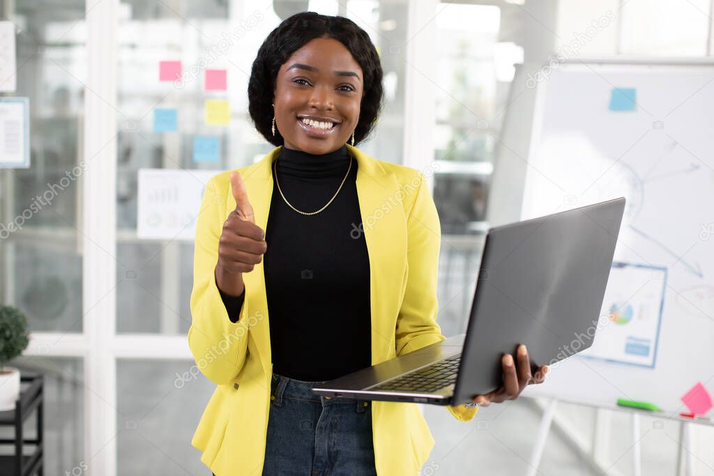 Smiling afro woman holding laptop while posing at office showing thumb up