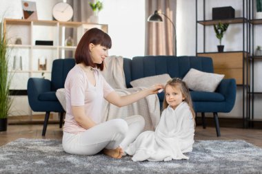 Güzel, genç bir anne banyo yaptıktan sonra küçük kızının saçını tarıyor. Havluya sarılı şirin bir kız çocuğu, annesiyle birlikte gri halıda oturuyor. Evdeki aydınlık oturma odasında yerde.