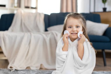 Bebek hijyeni ve cilt bakımı konsepti. Kapalı alanda, banyodan sonra havluya sarılı, evde yerde oturan ve iki pamuk diski yanaklarının yakınında tutan küçük bir kız çocuğu.