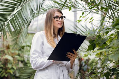 Bitki bakımı ve koruma konsepti. Serada çalışmak için. Hoş odaklı kadın tarım mühendisi seradaki güzel palmiye ağaçlarının önünde duruyor ve notlar alıyor.