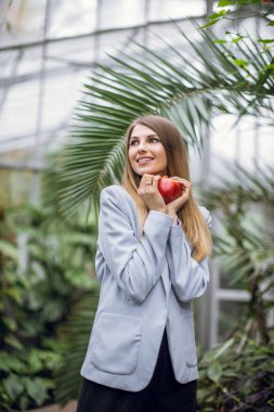 Yaz, insanlar, sağlıklı yaşam tarzı konsepti. Gülümseyen, açık gri ceketli mutlu sarışın kadın ellerinde kırmızı narı tutuyor ve uzaklara bakıyor, serada tropik bitkilerle birlikte poz veriyor.