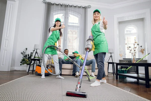 Two pretty women cleaning room with vacuum, while two multiracial men using tablet on sofa on the background