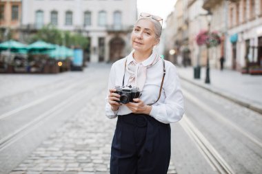 Elinde retro kamerayla dışarıda poz veren olgun bir kadın.