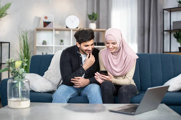 People, family and technologies concept. Happy young arabian muslim couple using app or social media on mobile phone at home, sitting on blue sofa in living room