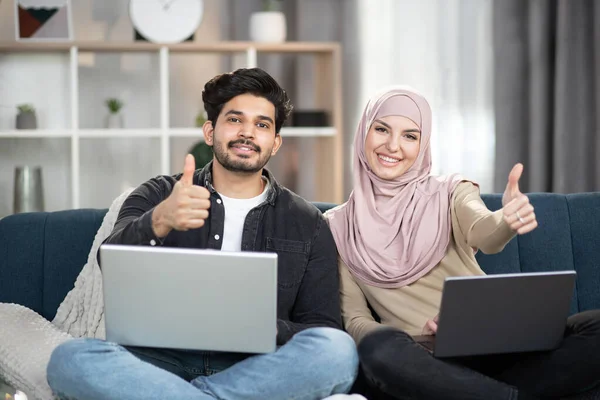 Jovem casal muçulmano multirracial relaxando no sofá em casa usando laptops, sorrindo para a câmera e mostrando os polegares para cima — Fotografia de Stock