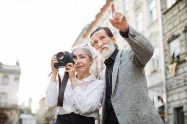 Yaşlı kadın kocası yakınlarda konuşurken fotoğraf çekiyor.