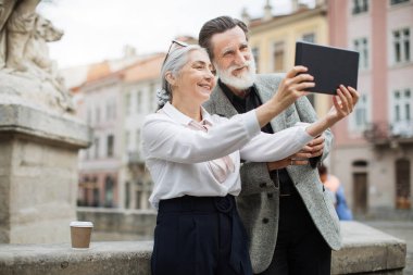 Gülümseyen yaşlı çift dışarıda dijital tabletle selfie çekiyor.