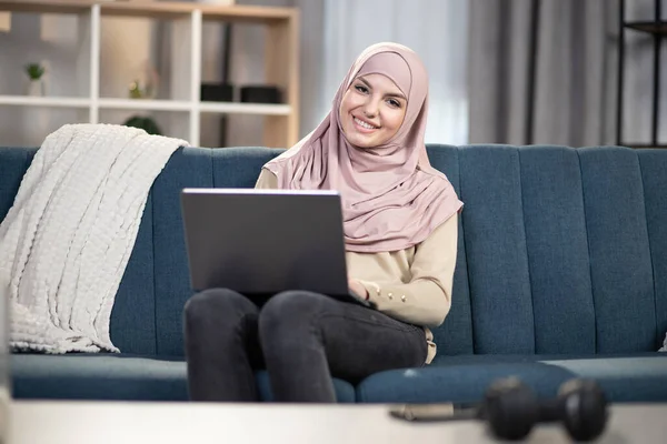 Distance work and freelance concept. Smiling beauty Muslim woman in hijab working remotely on laptop at home, sitting on sofa in living room. Online work and education